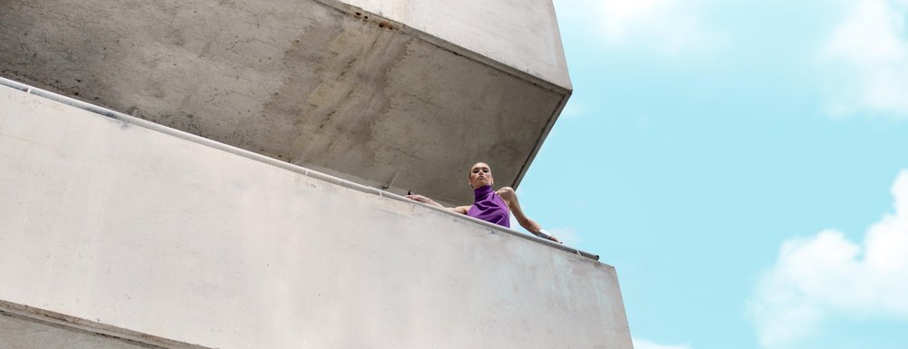 Model on the balcony of a concrete building, shot from below against the sky